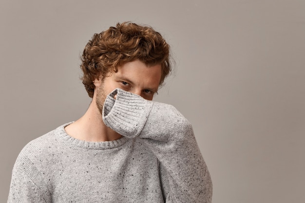 Body language and human facial expressions. trendy looking cunning young bearded guy wearing gray warm pullover, covering nose and mouth with hand, his look full of mystery