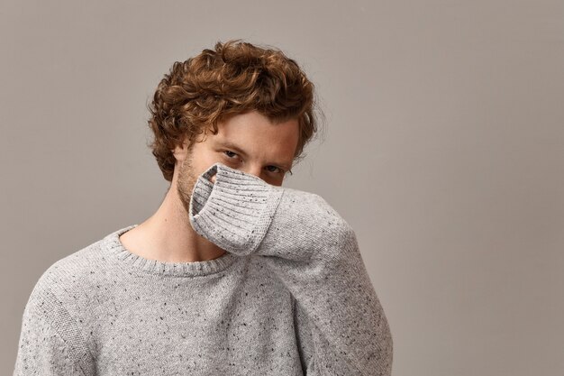 Body language and human facial expressions. trendy looking cunning young bearded guy wearing gray warm pullover, covering nose and mouth with hand, his look full of mystery