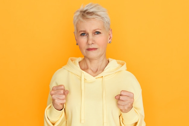 Body language. Isolated image of frustrated upset retired female with blonde short hairstyle clenching fists, ready to fight, showing her strength, posing at yellow studio wall wearing hoodie