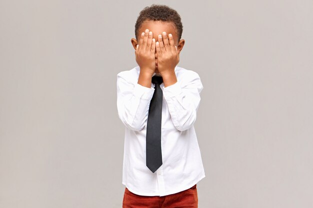 Body language. Isolated image of upset frustrated dark skinned male elementary pupil covering eyes with both hands, hiding his emotions, crying because of bad mark at school