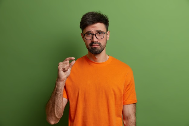 ボディーランゲージとサイズの概念。若い不機嫌な男は小さな測定を示し、彼の小さな給料について話し、何か小さなものを形作り、質の悪いものを判断し、オレンジ色のTシャツを着て、緑の壁に隔離されています