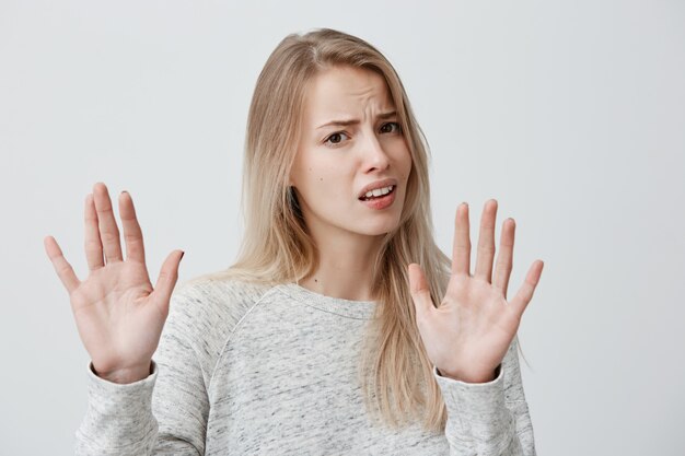 Body language. Stressed out woman wearing her blonde hair loose posing against gray wall, keeping hands in stop gesture, trying to defend herself as if saying: Stop that, Stay away from me