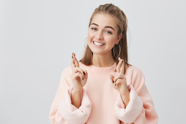 Body language. Superstitious teenager girl with blonde hair and pretty face crossing fingers for good luck, hoping her wishes will come true, having excited happy look, wearing pink sweatshirt.