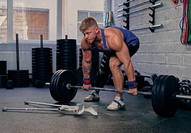 Bodybuilder with injured leg in bandage holds barbell near cross fit stand in a gym club.