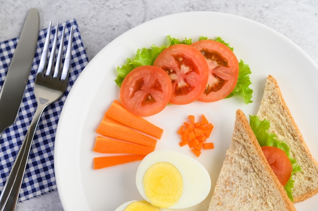 Boiled eggs, bread, carrots, and tomatoes on a white plate with a knife and fork.