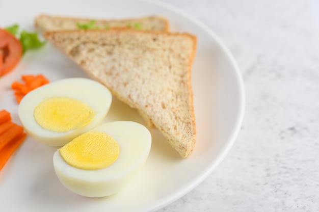 Boiled eggs, bread, carrots, and tomatoes on a white plate with a knife and fork.