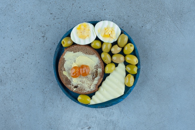 Boiled eggs, cheese slice, butterbrot and green olives on a blue platter on marble.