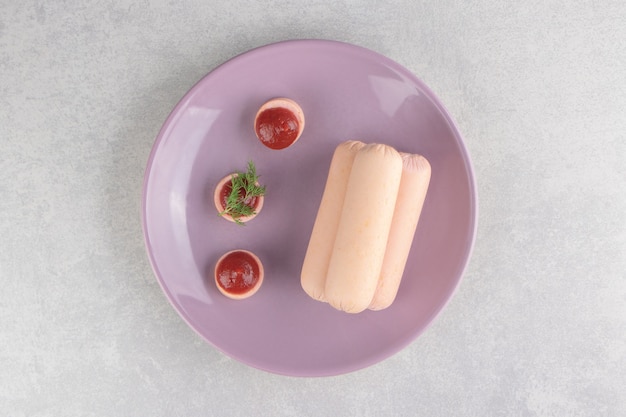 Вареные домашние сосиски и кетчуп на фиолетовой тарелке.