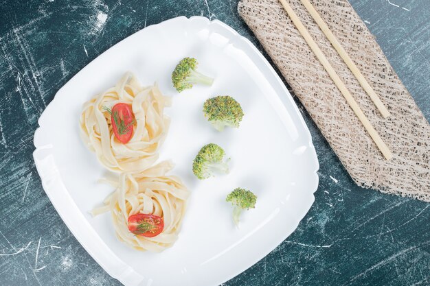 Вареные макароны тальятелле на белой тарелке с ломтиками брокколи и помидора. Фото высокого качества