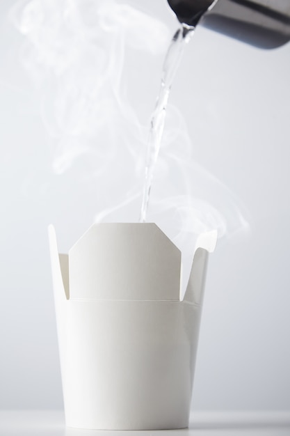 кипяток льется из чайника из нержавеющей стали в белый картонный контейнер для рамэн, изолированный на белом