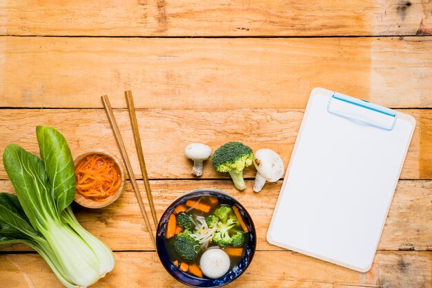 Bokchoy; carrot; fish ball soup; chopsticks and white blank clipboard over the table