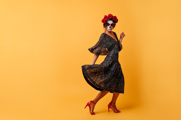 Bold dancing girl with dark hair stabbed with wreath of natural flowers on her head moves, posing with black dress and zombie mask