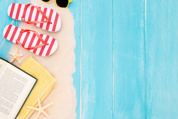 book, beach towel, flip flop, sunglasses, sand on blue wooden background