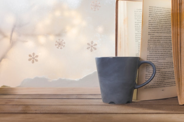 Book and cup on wood table near bank of snow, snowflakes and fairy lights