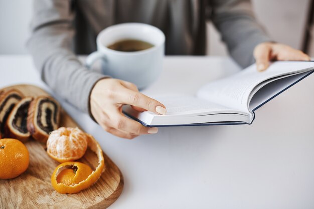 Книга как камера знаний. Шикарная современная женщина читает любимый роман во время завтрака, наслаждаясь горячим чаем в спокойной и уютной обстановке, пилинг мандарина и еды
