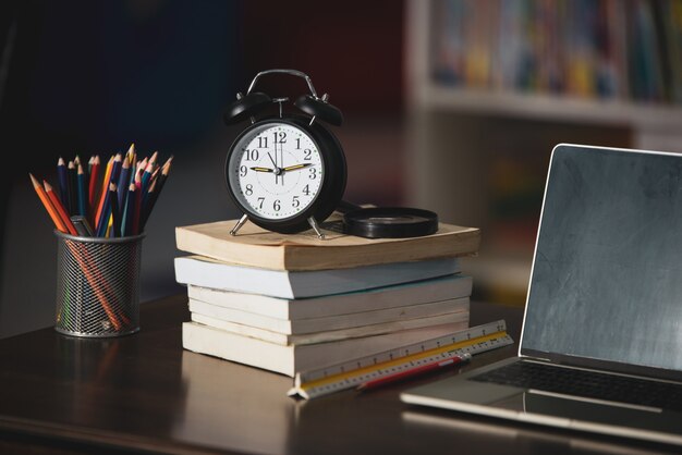 Book, laptop, pencil, clock on wooden table in library, education learning concept