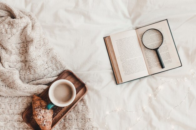 Book with loupe near croissant and hot drink on stand placed on plaid