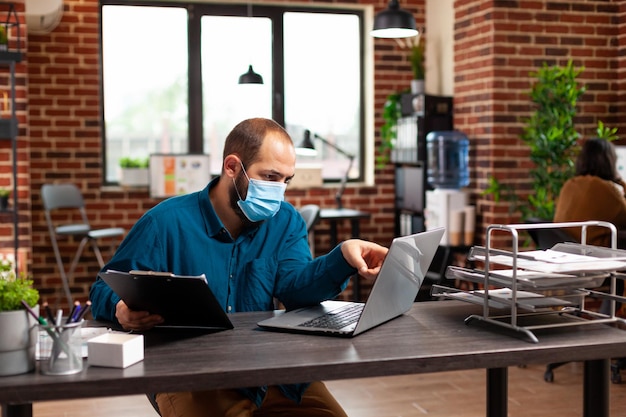 Bookkeeper man with medical protective face mask against coronavirus working at management strategy analyzing company report in startup office. Businessman planning business investment
