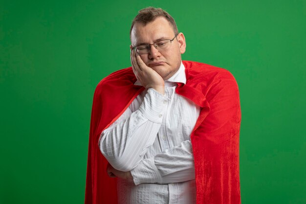 Bored adult superhero man in red cape wearing glasses putting hand on face looking straight isolated on green wall