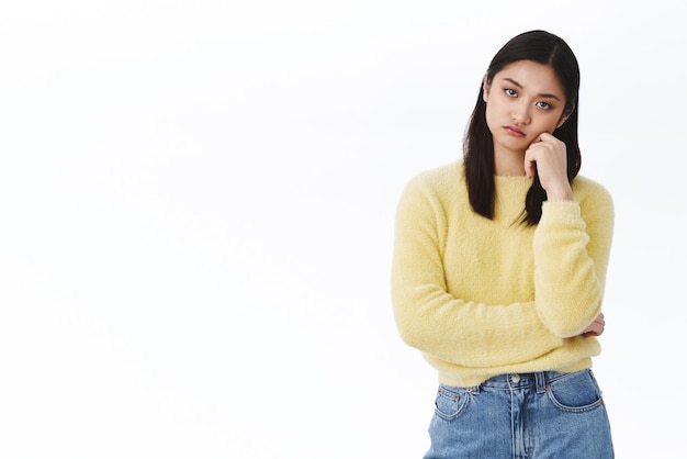 Bored asian girl looking at camera Skeptical and ignorant young woman in yellow sweater listening boring stories lean face on palm and staring without interest white background