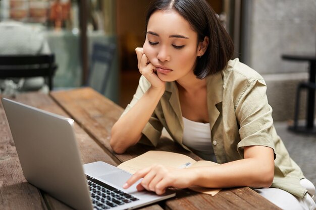 Скучающая азиатка сидит в кафе и смотрит на ноутбук, учится и грустит, делая домашнее задание