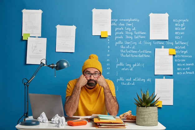 L'uomo barbuto annoiato lavora con il dispositivo portatile e le carte, tiene le guance, guarda la telecamera, si prepara per il seminario di studio