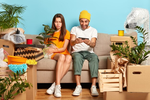 Скучающая европейка держит горшок с зеленым комнатным растением, смотрит в сторону на дисплей смартфона, наблюдает, как парень играет в онлайн-игры, вместе переезжает в недавно купленную квартиру