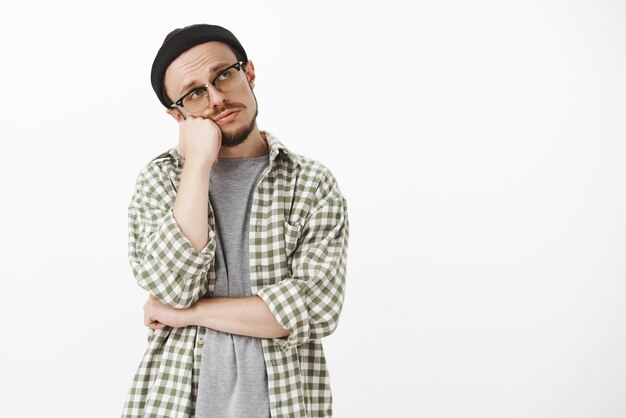 Bored frustrated gloomy handsome bearded guy in glasses black beanie and checked green shirt leaning head on fist gazing at upper right corner with lonely indifferent look expressing apathy