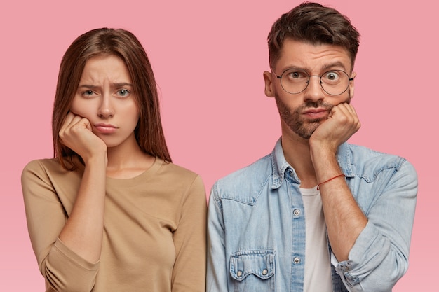 Bored girl and boy keep hand under chin, look with sadness directly, feel dissatisfied, wait for something for long time, stand against pink wall. People and facial expressions concept