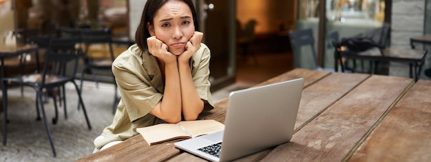 Foto gratuita una ragazza annoiata guarda il suo portatile seduta all'aperto in un bar ascoltando una noiosa riunione online di lavoro e