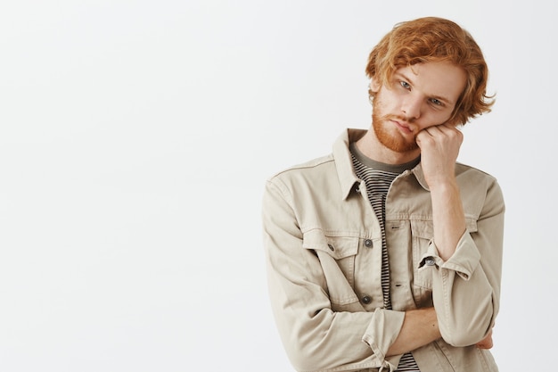 Bored and gloomy bearded redhead guy posing against the white wall