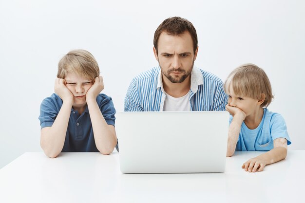 Bored indifferent son sitting with sibling and father at table, leaning head on hands while looking at laptop screen, checking grades in school