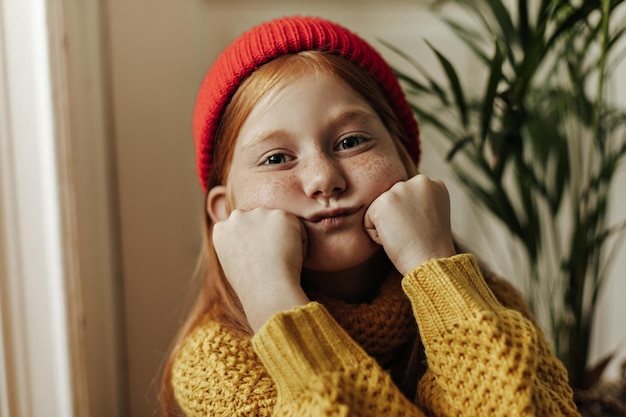 Скучающая маленькая девочка с рыжей прической и милыми веснушками в стильной кепке и желтом свитере смотрит вперед в квартире