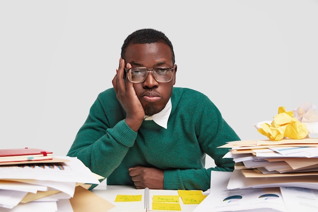 Bored male worker has tired facial expression, looks in displeasure, works long hours on project task, has dark healthy skin, sticks notes in notepad