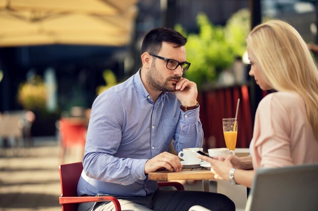 Bored man looking with suspicion at his girlfriend who is text messaging on cell phone and ignoring him in a cafe