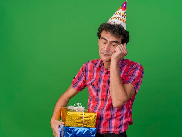 Bored middle-aged party man wearing birthday cap holding gift packs putting hand on face with closed eyes isolated on green wall