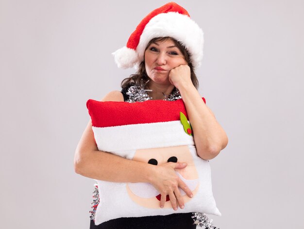 Bored middle-aged woman wearing santa hat and tinsel garland around neck holding santa claus pillow looking at camera keeping hand on face puffing cheeks isolated on white background with copy space