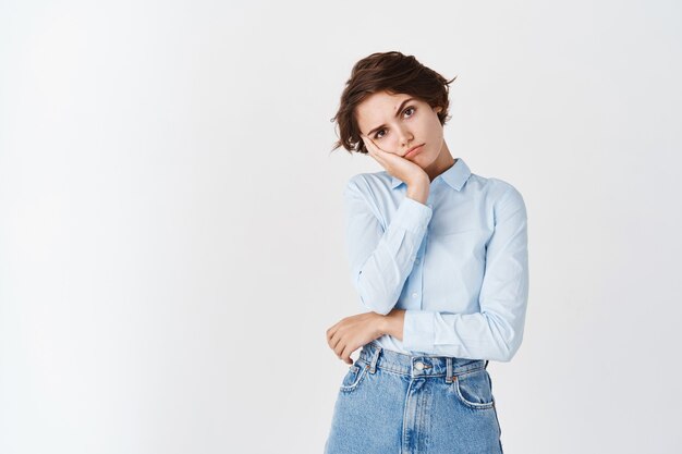 Bored and sad young caucasian woman in office clothing, lean face on palm and staring distressed  standing upset on white wall