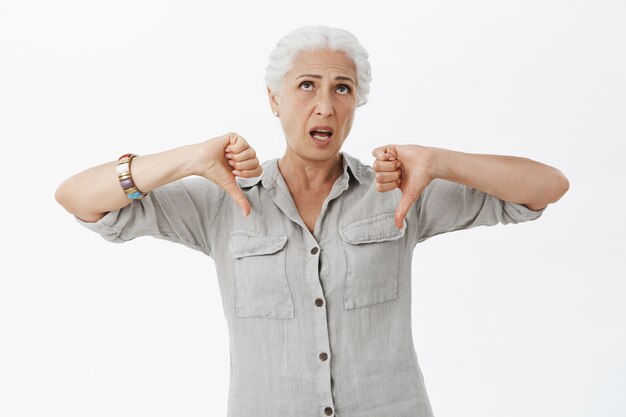 Bored skeptical elderly woman looking displeased, showing thumbs-down in dislike and roll eyes up