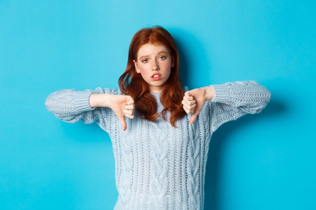 Bored and skeptical redhead girl showing thumbs down, looking unamused and uninterested, standing over blue background.