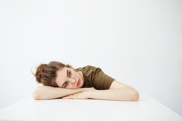 Bored tired dull young woman student with bun sitting at table over white background.