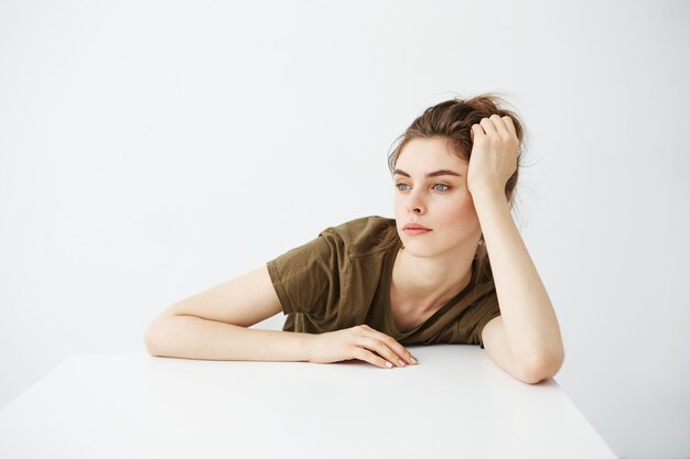 Bored tired dull young woman student with bun sitting at table over white background.