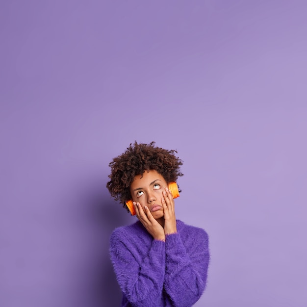 Free photo bored tiresome afro american woman keeps hands on cheeks focused above wears headphones to listen favorite music dressed in casual sweater