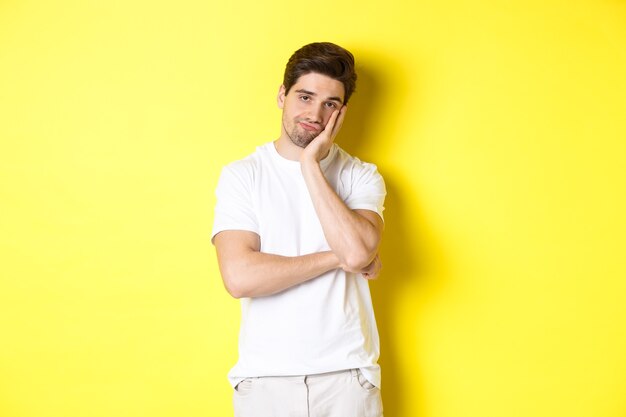 Bored and unamused man looking with indifference, standing in white clothes over yellow background