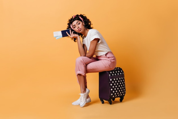 Скучная женщина в розовых штанах с билетами на самолет студийный снимок модной девушки, сидящей на чемодане