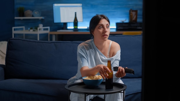 Bored woman relaxing on cozy couch, eating popcorn and drinking beer using remote controller switching tv channels, searching funny online film movie, spending free weekend time alone at home.
