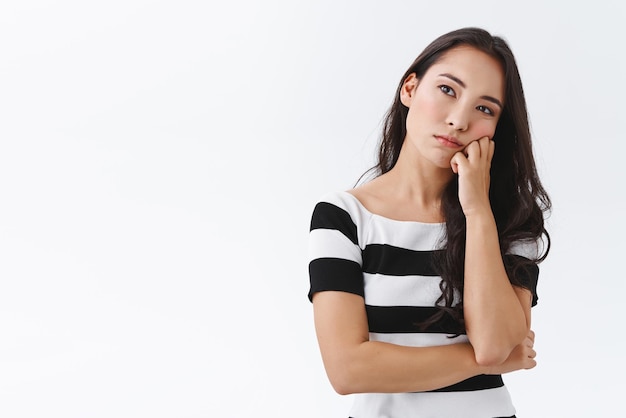 Annoiato giovane donna asiatica guardando in lontananza con distratta espressione triste faccia magra sul pugno tirare smorfia infastidito in piedi incontro disinteressato su sfondo bianco pensando pensieroso