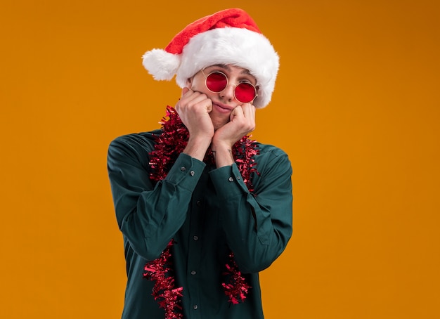 Bored young blonde man wearing santa hat and glasses with tinsel garland around neck looking at camera keeping hands on chin isolated on orange background with copy space