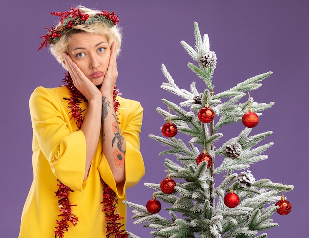 Bored young blonde woman wearing christmas head wreath and tinsel garland around neck standing near decorated christmas tree looking  keeping hands on face isolated on purple wall