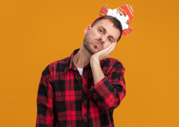 Bored young caucasian man wearing santa claus headband looking at camera keeping hand on face with pursed lips isolated on orange background with copy space